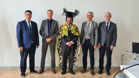 Männliche Personen stehen nebeneinander und lächeln in die Kamera. Der Mann in der Mitte ist mit einem Doktorhut gekennzeichnet.