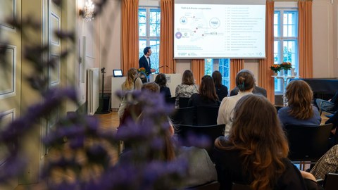 A male person with black hair is giving a presentation at the lectern (left). The presentation area is on the right.