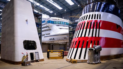 The halls of a shipyard. In the center: a large ship, with other large shipbuilding components to the left and right.