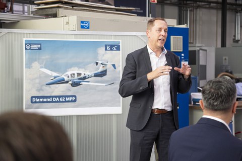 A man in a suit with brown hair speaks passionately during a lecture. In the background is a poster featuring the research aircraft.