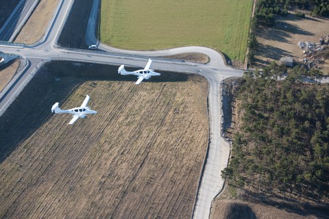 Flugaufnahme zweier Flugzeuge von der Seite aus größerer Distanz. Im Hintergrund befindet sich Ackerland.