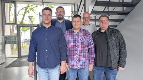 Fünf Männer stehen in einem Foyer an einer Treppe und schauen grinsend in die Kamera.