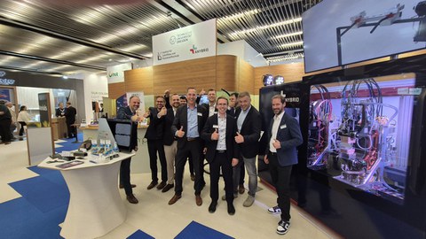 A group of men are standing together and looking at the camera. They are all laughing and giving a thumbs up. They are at a trade fair booth. Around them are machines and demonstrators on tables.