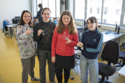 A group of girls stand in a semi-circle and smile at the camera. They are holding homemade bottle openers and key rings.