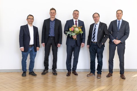 Five men in suits stand in a row and smile at the camera. The person in the middle is holding a bouquet of flowers.
