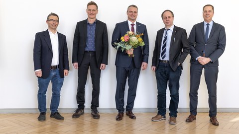 Five men in suits stand in a row and smile at the camera. The person in the middle is holding a bouquet of flowers.