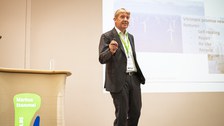 Ein Mann in einem schwarzen Anzug hält einen Vortrag. Neben ihm steht ein Pult mit seinem Namen, hinter ihm werden Folien an die Wand projiziert. Er ist sehr dynamisch.