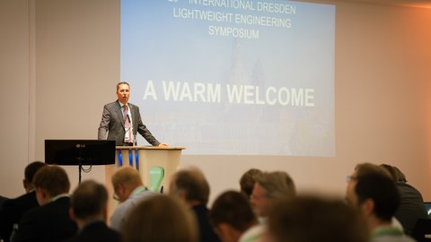 Ein Mann steht an einem Rednerpult und redet. Im Vordergrund sitzen viele Menschen, die ihm zuhören. Man sieht sie von hinten.