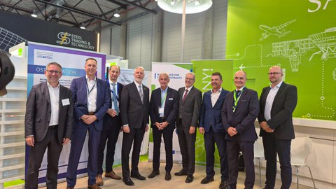Group photo: Nine men are standing side by side at a booth at the ILA Berlin, smiling at the camera.