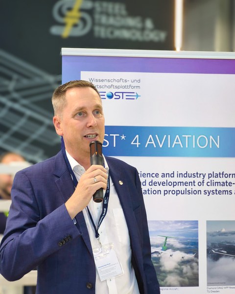 Prof. Gude, ein Mann in einem Anzug, redet in ein Mikrofon. Hinter ihm steht ein OST4Aviation Roll-up.