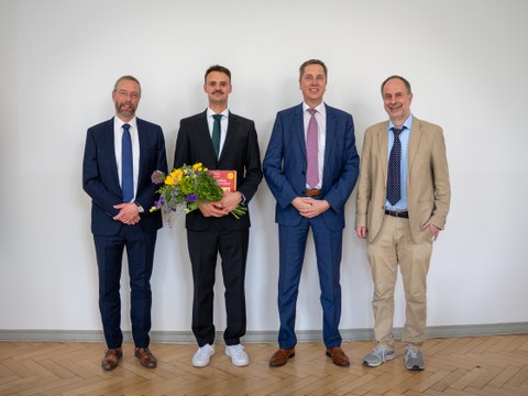 Vier Männer im Anzug, der zweite von links mit einem Strauß Blumen in der Hand, stehen nebeneinander und lächeln in die Kamera.