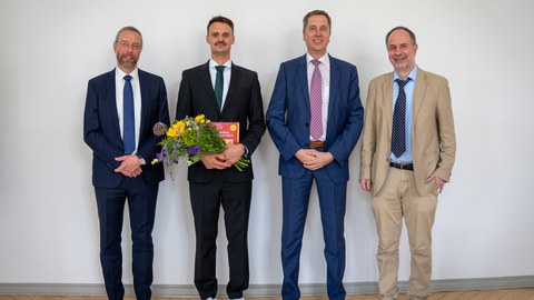 Vier Männer im Anzug, der zweite von links mit einem Strauß Blumen in der Hand, stehen nebeneinander und lächeln in die Kamera.