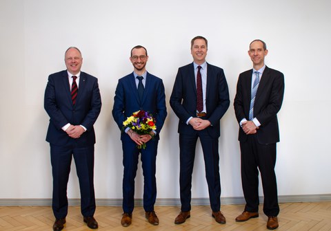 Four men in suits are standing in a row and smiling at the camera. The second person from the left is holding a bouquet of flowers.
