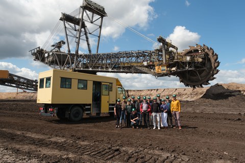 A group of people stands in the open-cast mine area. A mining vehicle can be seen in the background.