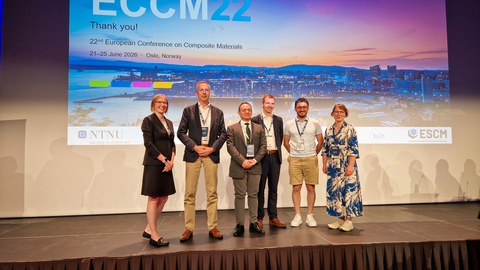 Six people are standing on a stage, looking at the camera. An ECCM-22 slide is being projected behind them.