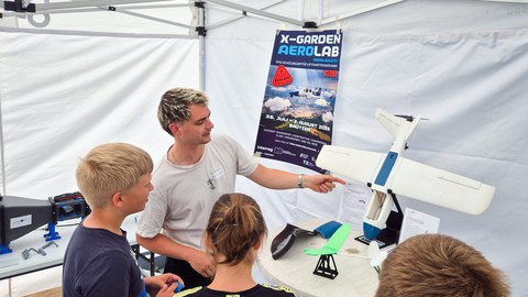 A young engineer stands inside the pavilion, pointing to a model aircraft while children stand around and listen.