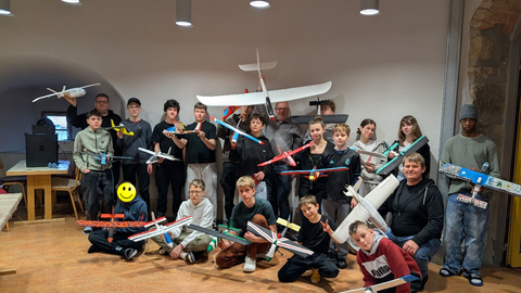 Group photo of students and supervising engineers at the X-Garden AeroLab. The participants are smiling at the camera and presenting the model aircraft they designed and built themselves.