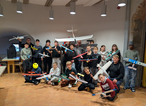 Gruppenbild von Schüler:innen und betreuenden Ingenieur:innen beim X-Garden AeroLab. Die Teilnehmenden lächeln in die Kamera und präsentieren ihre selbst konstruierten und gebauten Modellflugzeuge.