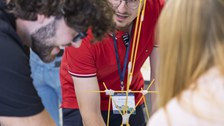 Im Vordergrund ist ein Spaghettiturm zu sehen, der durch Klebeband verbunden ist. Im Hintergrund ist ein Student zu sehen, der sich über den Tisch lehnt.