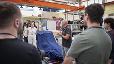 Student:innen stehen im Halbkreis und schauen einen glatzköpfigen Mann an, welcher etwas erzählt. Sie befinden sich in einer Produktionshalle o.ä.