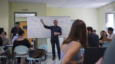 Ein Mann steht vor einer Tafel und erklärt den Student:innen, die drum herum sitzen, gestikulierend etwas.