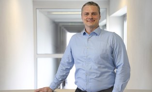 Ein Mann steht in der Mitte eines Raums und stützt sich ab. Er lächelt in die Kamera. Es ist eine Portraitaufnahme. 