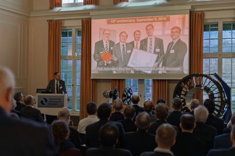 Ein Mann steht an einem Pult, auf einer Leinwand ist ein Gruppenbild von vier Männern projiziert. Darüber findet man die Überschrift "10th anniversary, September 8, 2016". Publikum schaut auf die Szene vor sich. In der rechten Ecke stehen Demonstratoren.