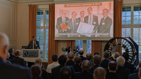 Ein Mann steht an einem Pult, auf einer Leinwand ist ein Gruppenbild von vier Männern projiziert. Darüber findet man die Überschrift "10th anniversary, September 8, 2016". Publikum schaut auf die Szene vor sich. In der rechten Ecke stehen Demonstratoren.