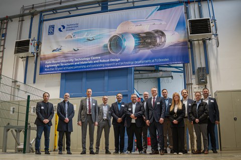 Menschen in Businesskleidung stehen aufgereiht unter einem Banner in technischer Umgebung. Auf dem Banner ist ein futuristisches Flugzeug abgebildet mit der Aufschrift "More than 20 years of trusted partnership and outstanding research and technology..".