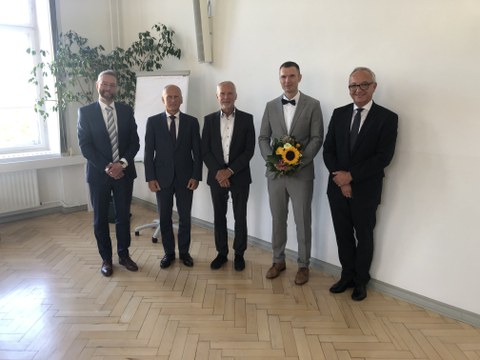 Foto von 5 Personen in Businesskleidung, die in die Kamera schauen. Eine Person hält einen Blumenstrauß.