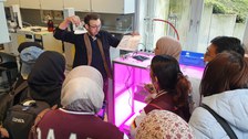 Ein Mann zeigt Studierenden einen Erlenmayerkolben mit rosa Flüssigkeit.
