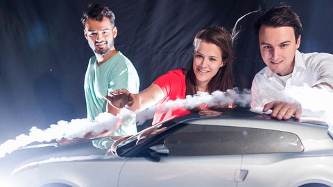 Foto: 3 Studierende blicken begeistert auf ein Modellauto, das von einer sichtbaren Luftströmung umflossen wird. 