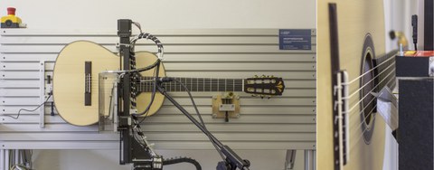 Das Foto ist zweigeteilt. Der überwiegende linke Teil zeigt eine Akustikgitarre, die in eine Wandvorrichtung gespannt ist. Der Anzupftest untersucht wie lange es dauert, bis ein Ton hörbar ist und wie lange der Ton hörbar bleibt. Der kleine rechte Teil des Fotos zeigt eine Detailaufnahme der Gitarrensaiten, die gerade von der Vorrichtung angezupft werden.