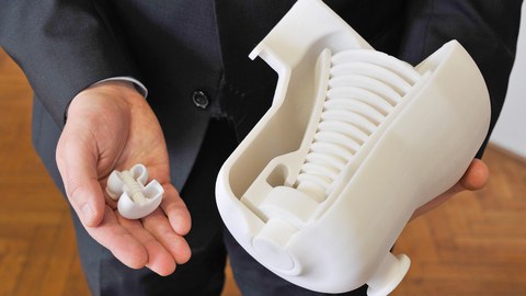 Das Foto zeigt die Nahaufnahme von zwei männlichen Händen. In der einen Hand hält er eine sehr kleine 3D-gedruckte Turbine. In der anderen Hand hält er eine etwa zehnmal größere 3D-gedruckte Turbine.