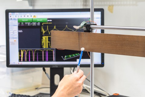 Ein Brett ist horizontal in eine Halterung eingespannt. Man sieht eine Hand, die einen kleinen Hammer hält, der gerade auf das Holz klopft. Im Hintergrund steht ein Monitor, der verschiedene mehrfarbige Kurven zeigt. Damit testen die Wissenschaftler die Schwingungseigenschaften von thermisch behandeltem Ahorn. Diese sind entscheidend für die Klangqualität.