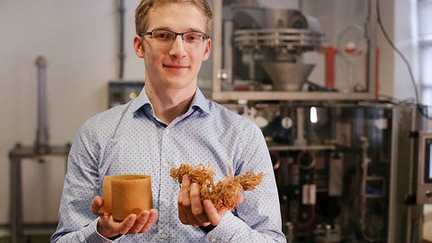 Das Foto zeigt den Nachwuchswissenschaftler Ludwig Schmidtchen, der in seinen Händen hellbraune, getrocknete Algen hält. Im Hintergrund ist eine Laborapparatur zu sehen.