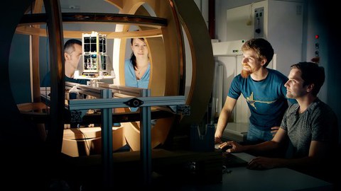 4 Studierende und Mitarbeitende der TU Dresden sind im Labor bei Tests und betrachten den in der Mitte aufgebauten Satelliten