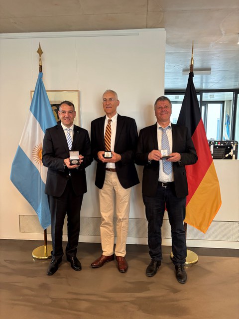 Three middle-aged men in suits pose with their prizes. In the background are the Argentinian and German flags.
