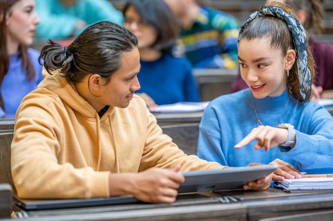  Bilder von zwei Studierende, die in einem vollen Hörsaal nebeneinander sitzen und sich gegenseitig helfen.