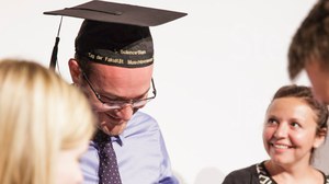 Foto: ein Mann mit Doktorhut blickt nach unten und betrachtet etwas, das nicht zu sehen ist, 2 weitere Frauen sind zu sehen, eine der Frauen schaut ihn erwartungsvoll an
