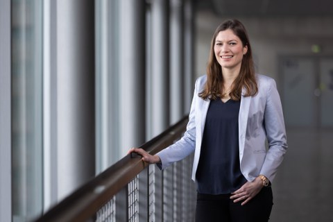 Anja Wagemans steht an ein Geländer gelehnt. Sie hat lange braune Haare, trägt einen hellen Blazer und lächelt.