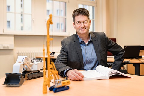 Foto von Professor Frank Will an einem Tisch auf dem 2 Modelle von Baumaschinen stehen