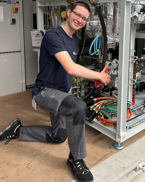 Junger Mann mit kurzen Haaren und Brille in Arbeitskleidung hantiert mit einer Zange an einer Mutter in einer Werkstatt. Er lächelt in die Kamera.