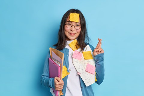 Asiatische Frau kreuzt Finger. Sie hält Ordner und ist mit Post-Its beklebt. Sie ist in der Prüfungsvorbereitung.