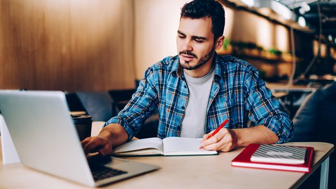 Das Foto zeigt einen jungen Mann, der an einem Tisch sitzt. Während er mit einer Hand auf seinem Laptop tippt, schreibt er mit der anderen etwas in ein Notizbuch.