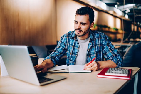 Das Foto zeigt einen jungen Mann, der an einem Tisch sitzt. Während er mit einer Hand auf seinem Laptop tippt, schreibt er mit der anderen etwas in ein Notizbuch.