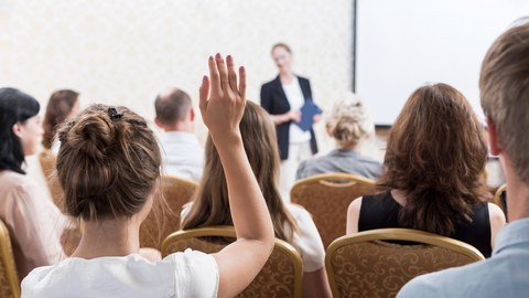 Foto von Zuhörern in einem Seminar. Die Zuhörer sind mit dem Rücken dem Betrachter zugewandt und einer von ihnen meldet sich. 
