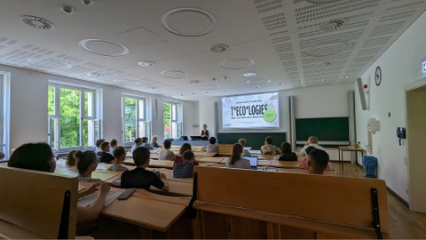 Personen sitzen im Hörsaal und schauen auf eine Projektion.