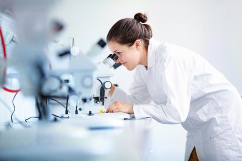 Auf dem Foto sieht man eine junge Wissenschaftlerin im weißen Kittel, die durch ein Mikroskop schaut.