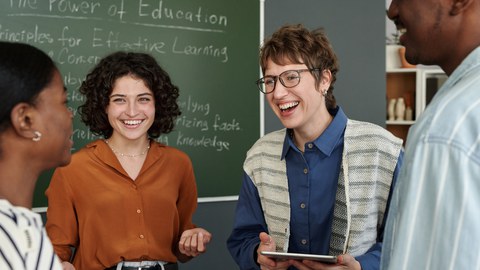 Gruppe von Personen, die in einem Klassenzimmer mit einer Kreidetafel im Hintergrund über Grundsätze für effektives Lernen diskutieren. Die Gruppenmitglieder lächeln und sind in ein Gespräch vertieft.